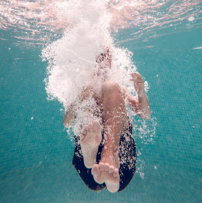 Underwater full body shot of unrecognizable barefoot kid wearing swimming shorts plunging in transparent clean blue water in pool